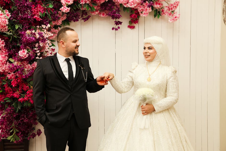 Bride and groom in elegant attire posed with vibrant floral wall backdrop, indoors.